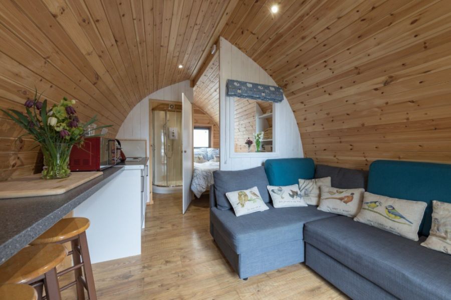 Glamping Pod with a light wood ceiling, a blue sofa, dining table and with a bed and bathroom at the back