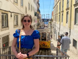 Chelsea, Founder of Cheap Holiday Expert in a blue shirt standing on a narrow Lisbon street, surrounded by tall buildings, pedestrians and an old yellow tram